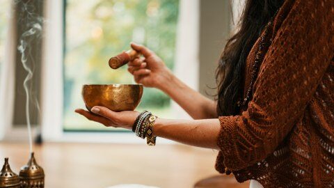 Eine Frau, deren Gesicht nicht zu sehen ist, steht vor einem Räucherstäbchen und hält eine Klangschale in der Hand