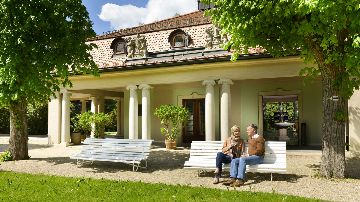 Ein Paar sitzt im Sonnenschein auf einer Bank vor der Wettinquelle in Bad Brambach