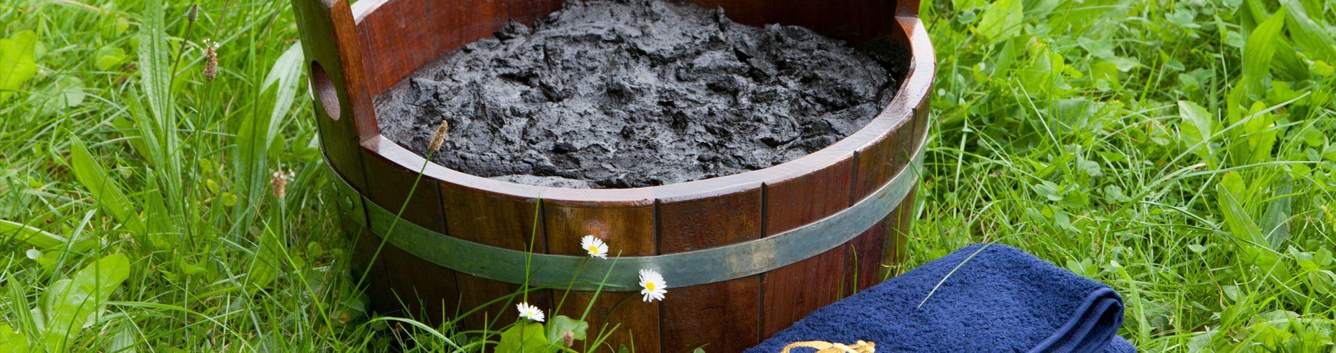 Ein Holzkübel gefüllt mit Naturmoor steht auf einer grünen Wiese und neben dran liegt ein blaues Handtuch