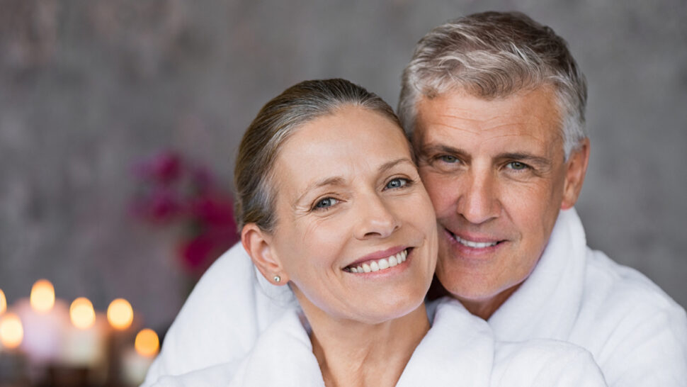 Ein Paar mittleren Alters haben weiße Bademäntel an und schauen lächelnd hintereinanderstehend