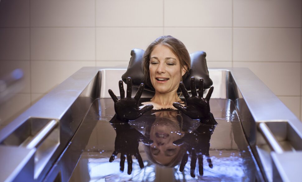 Eine Frau liegt in einer Naturmoorbadewanne und schaut lächelnd ihre Naturmoorhände an