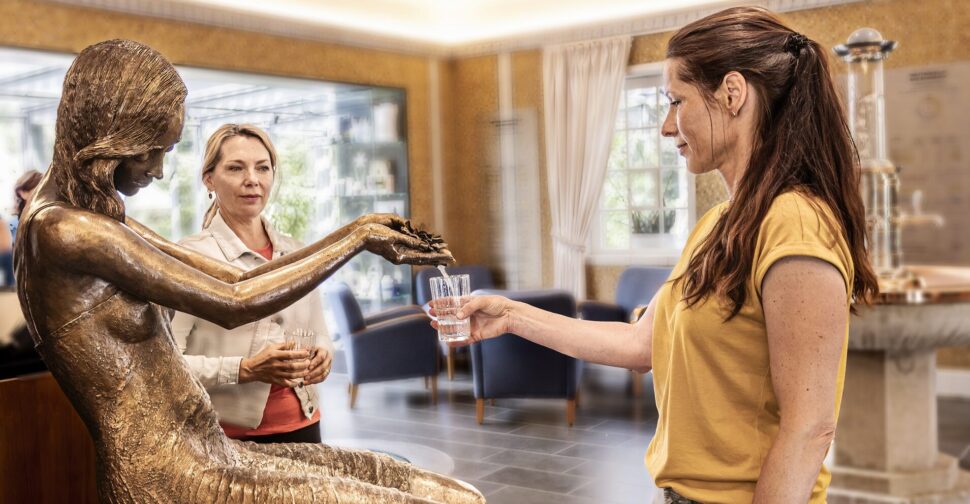 Zwei Frauen stehen vor der Wettinquellen-Statue und eine lässt sich das Glas mit Wasser voll laufen