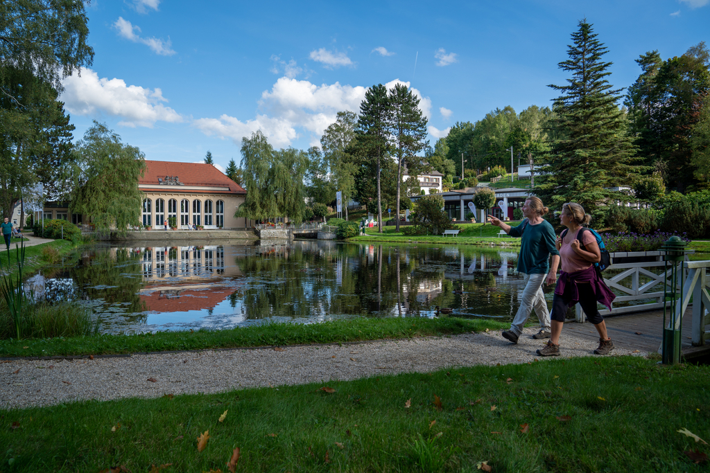 Zwei Personen laufen im Kurpark am Teich entlang, im Hintergrund ist die Festhalle zu sehen