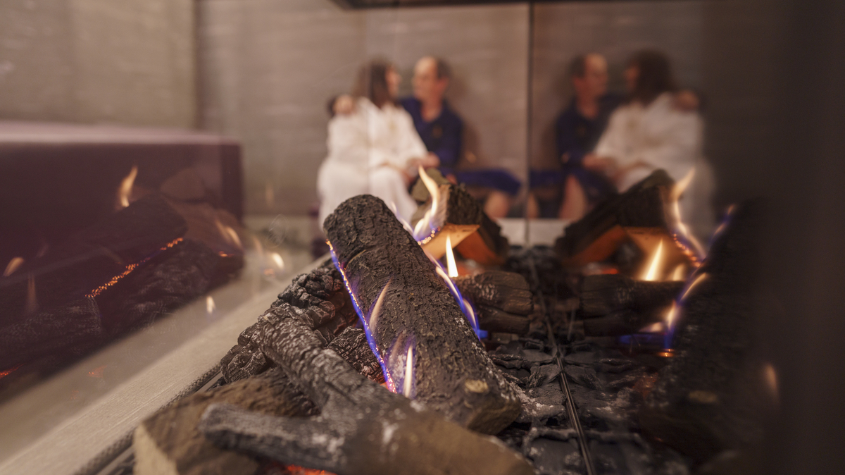 Blick auf ein brennendes Feuer in einem Ofen aus Glas, im Hintergrund ist unscharf ein Paar zu erkennen