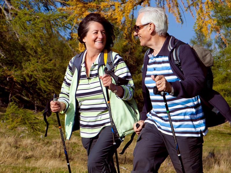 Ein älteres Paar mit Walkingstöcken läuft auf einem Feldweg und lächelt sich an