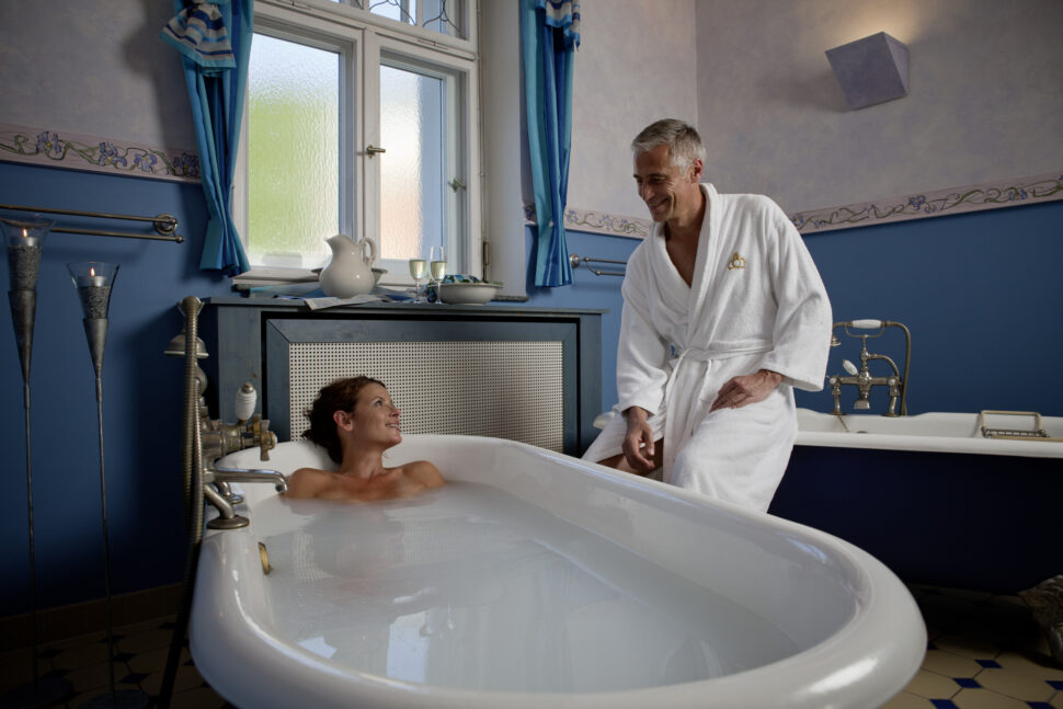 In einem Badewannenraum liegt eine Frau in milchigem Wasser und lächelt einen Mann an, der auf dem nächsten Badewannenrand sitzt